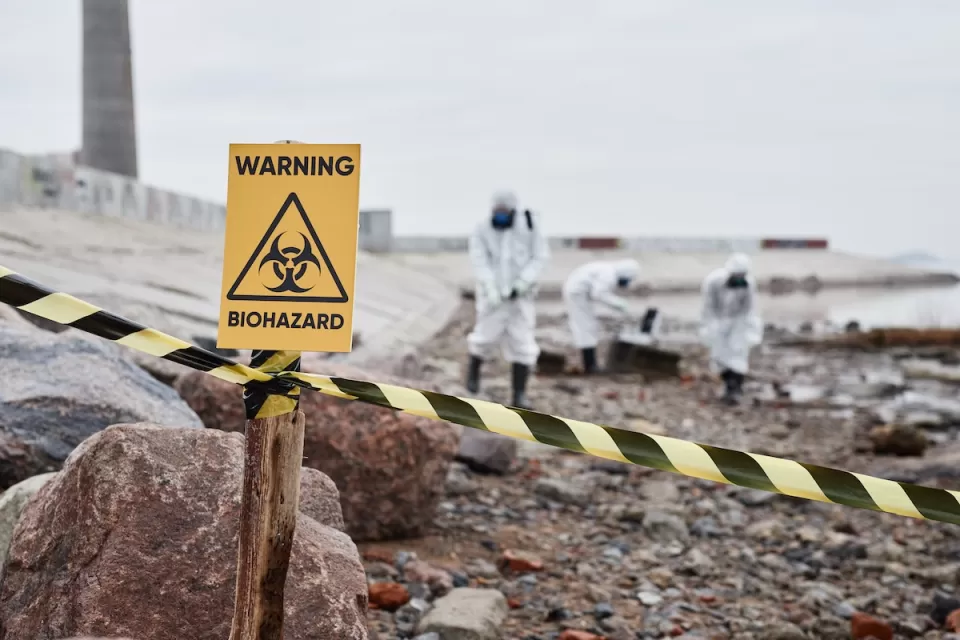 Close-up of yellow "Warning Biohazard" sign with people in hazmat suits cleaning up in background.